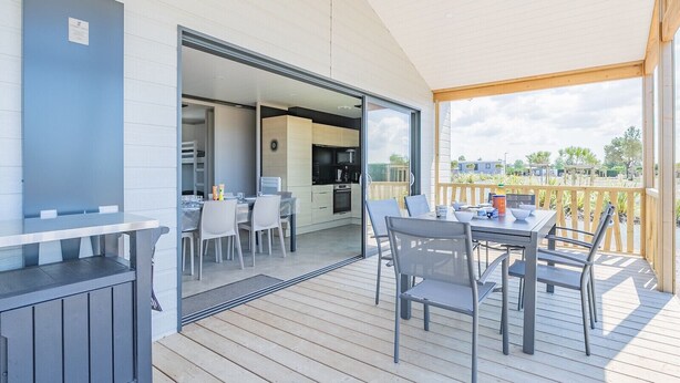Un área de comedor exterior moderna con terraza de madera, cocina con paredes de vidrio y vistas del paisaje circundante.