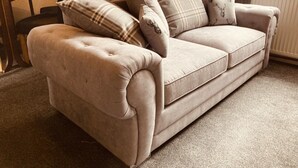 A beige sofa with a tufted backrest and armrests, accompanied by plaid pillows.