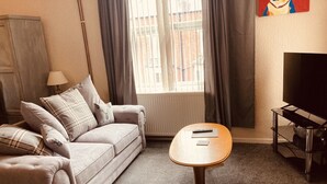 A living room with a sofa, a round wooden coffee table, a TV stand, and a window with curtains.