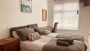 A bedroom with two beds, a desk, a chair, a window with blinds, and a wall-mounted mirror.