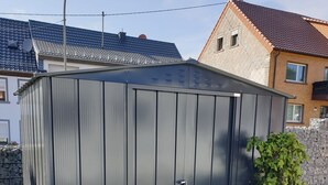 A metal shed with a sliding door, located in a grassy yard with a stone wall and a house in the background.
