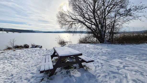 A snow-covered picnic table by a lake with bare trees and distant hills.
