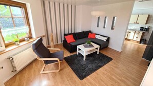 A modern living room with a black sofa, a white coffee table, and a black rug. There is a window with a view of greenery outside.