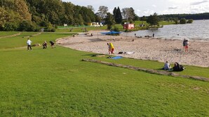 A lakeside park with grassy areas, a sandy beach, and a small red building.