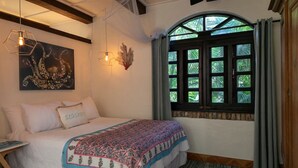 A bedroom with a large bed, a window with a view of greenery, a wall-mounted artwork, and a geometric light fixture. - One Bedroom Cottage on Peter Bay Beach (St John)