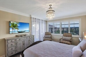A bedroom with a large bed, a television on the wall, a chandelier, and a window with a view of buildings.