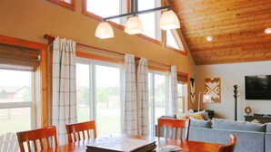 Une pièce spacieuse avec un plafond en bois, une table à manger avec des chaises, un canapé gris et de grandes fenêtres avec des rideaux.
