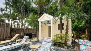 Un jardin avec des chaises longues, un petit abri de jardin et des plantes tropicales.