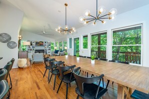 Un vaste espace de salle à manger avec une longue table en bois et des chaises noires, de grandes fenêtres donnant sur un paysage verdoyant et un lustre moderne.