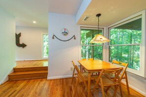 Un espace repas avec une table et des chaises en bois, une lampe suspendue et une vue sur la verdure grâce à de grandes fenêtres.