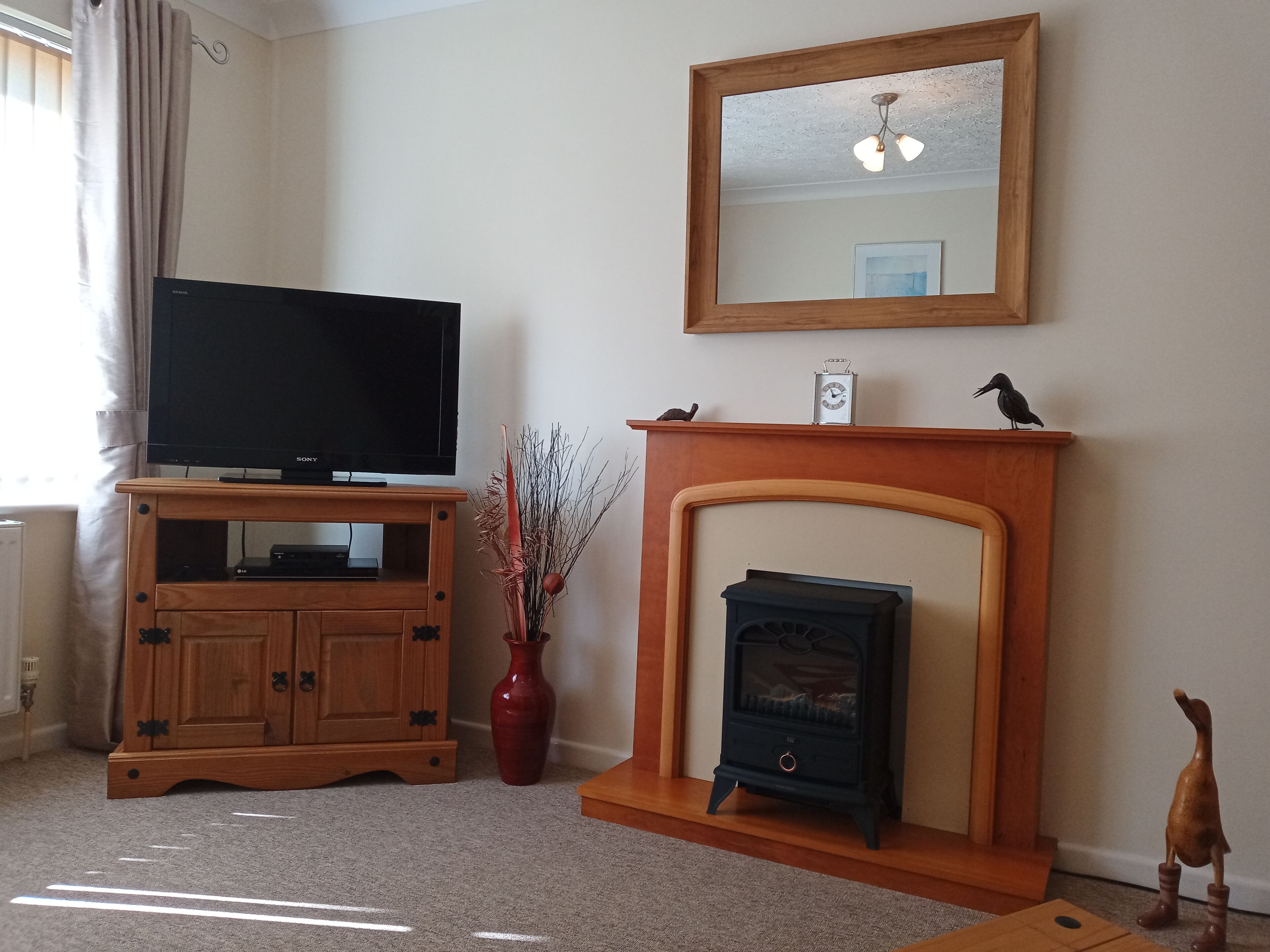 Una sala de estar con televisor sobre un mueble de madera, chimenea, espejo y un florero con ramas.
