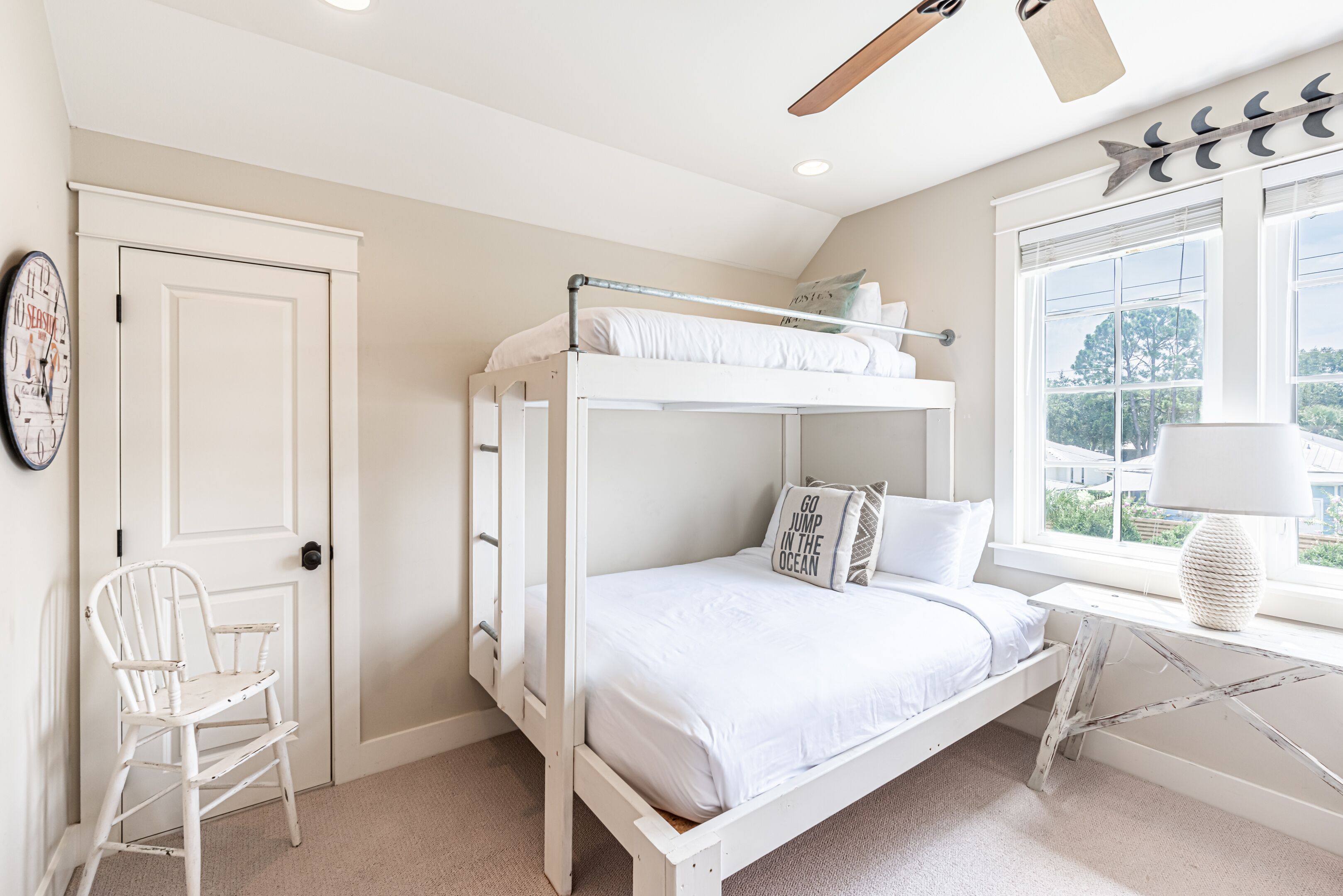 Un lit superposé avec une literie blanche, une chaise blanche, une fenêtre donnant sur un beau paysage et un ventilateur de plafond.