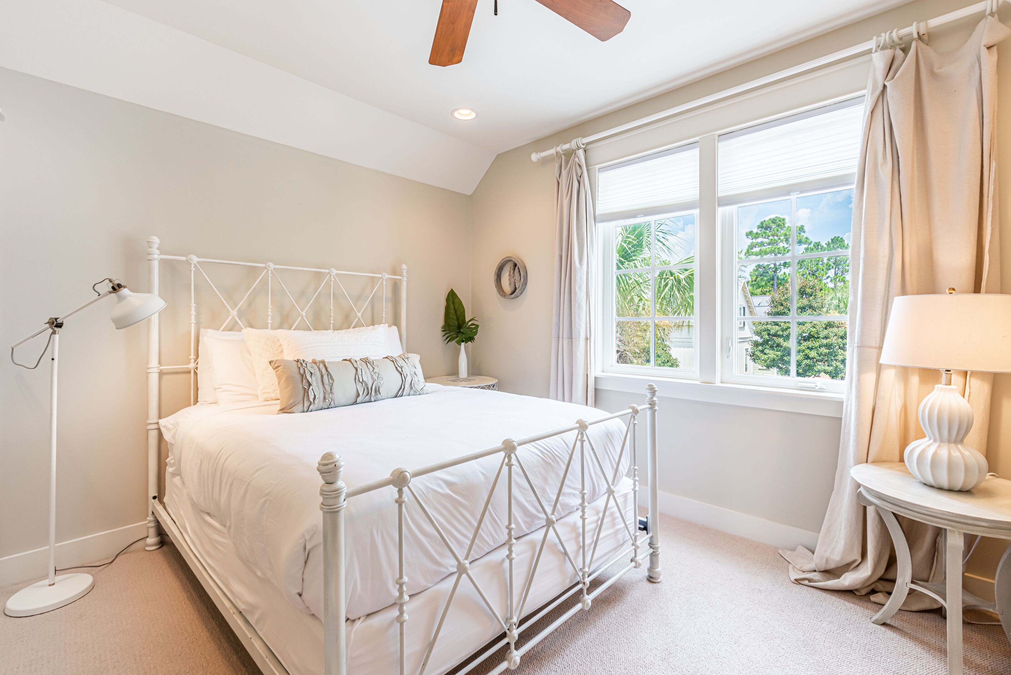 A bedroom with a white metal bed frame, a bedside lamp, a round table with a lamp, a window with curtains, and a plant on a side table.