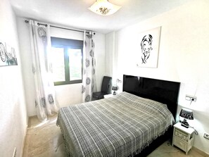 Een slaapkamer met een bed, een nachtkastje, een lamp, een raam met gordijnen en een muur met een schilderij.