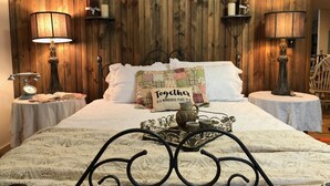 Quarto com parede de madeira, duas luminárias de cabeceira, cama com colcha, travesseiro decorativo e bandeja com bule.