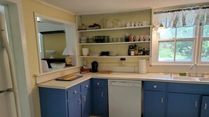 A compact kitchen with blue cabinets, a white dishwasher, and a window with a view of greenery. - Pine Island Retreat - 3BR's, 1st & 2nd level porches NO LINENS PROVIDED (Chincoteague)