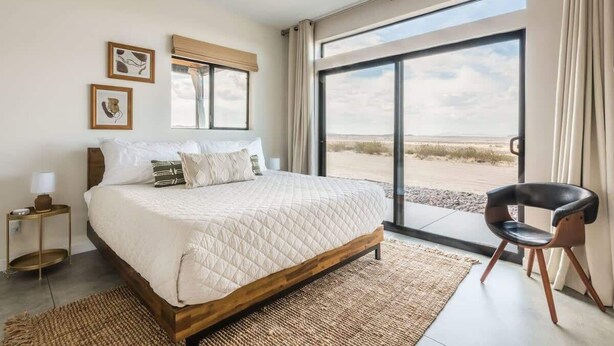 A bedroom with a large bed, a chair, a nightstand, and a view of the outdoors. - Estrella Perdida by Homestead Modern (Joshua Tree)