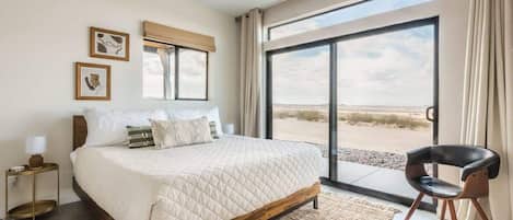 A bedroom with a large bed, a chair, a nightstand, and a view of the outdoors.