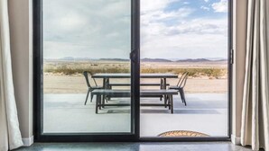 A sliding glass door leading to an outdoor patio with a table and chairs, a view of a desert landscape, and a clear sky. - Estrella Perdida by Homestead Modern (Joshua Tree)
