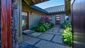 A modern house with a stone exterior, a wooden door, and a stone pathway leading to the entrance. - Luxury Home w/Pool + Spa | Sleeps 10 | Waikoloa Resort (Waikoloa)