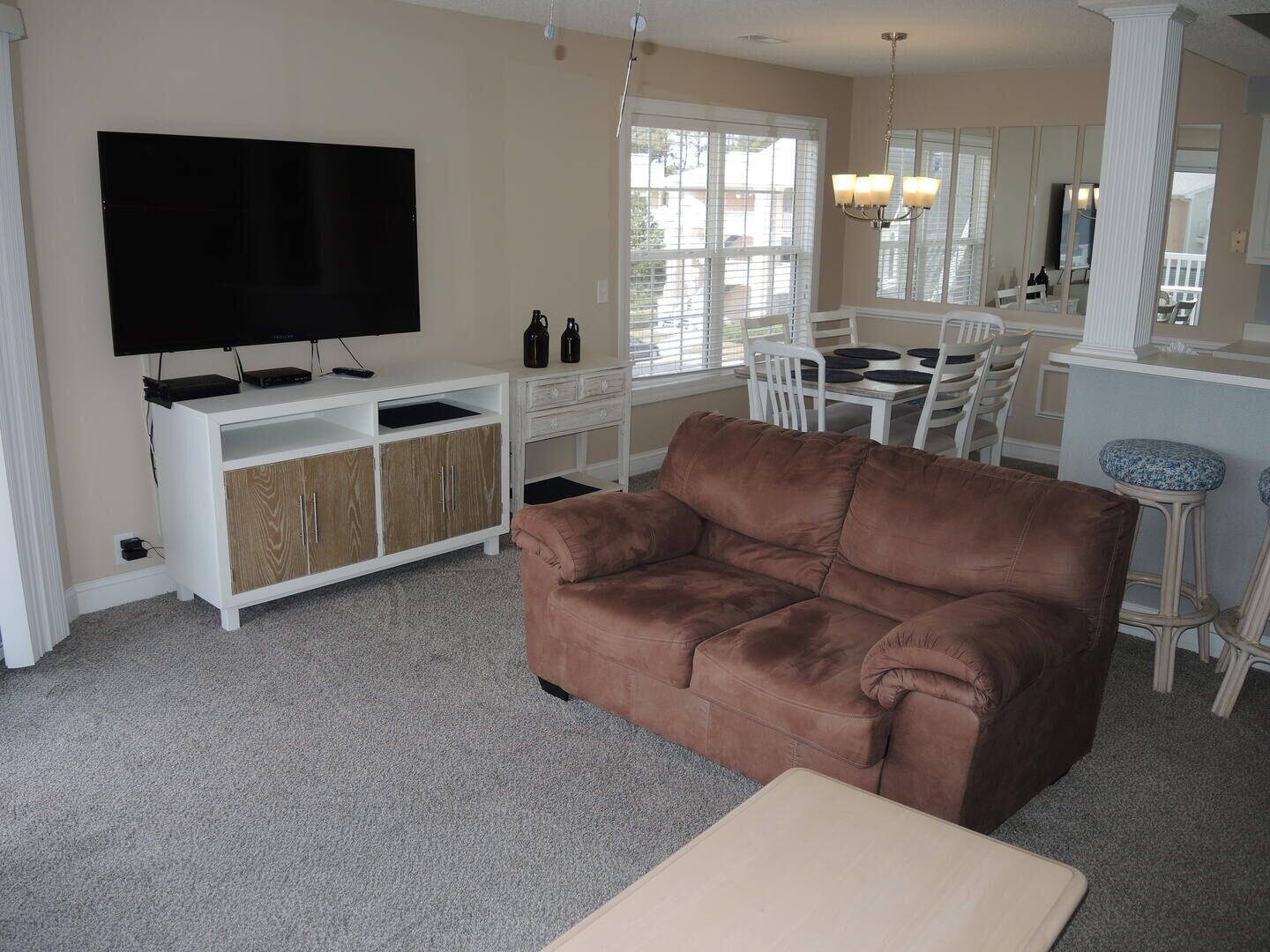 Ein Wohnzimmer mit braunem Sofa, einem Flachbildfernseher, einer weißen Unterhaltungsecke und einem Essbereich mit weißem Tisch und Stühlen.