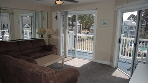 A living room with a brown sofa, a coffee table, and a ceiling fan. There is a sliding glass door leading to a balcony with outdoor furniture.