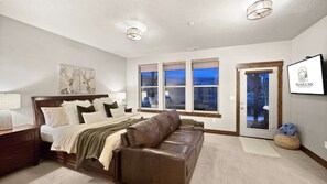 Een slaapkamer met een bed, een houten bankje, een nachtkastje, een lamp, een raam met uitzicht en een lijst met een afbeelding aan de muur.