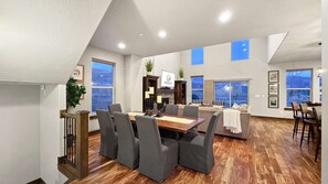 Een ruime woonkamer met een eettafel, stoelen, een open haard en grote ramen met uitzicht op de omgeving.