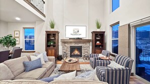 A spacious living room with a fireplace, comfortable seating, and a large window offering a view. - Fall Mountain Retreat•Hot Tub•BBQ•Community Amenities•Uinta Foothill Views (Heber City)