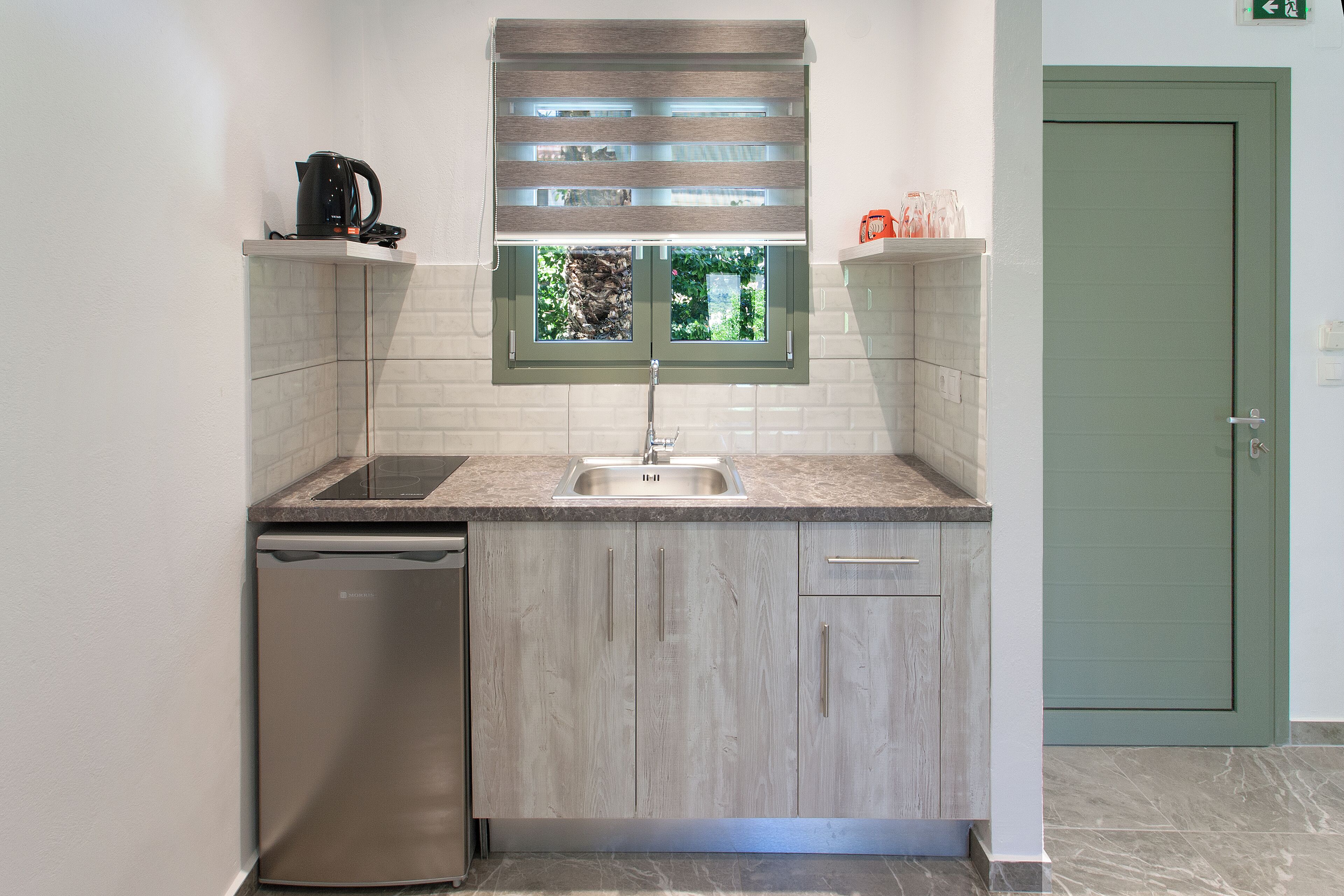 Una cocina compacta con lavavajillas integrado, una ventana con persianas y una puerta verde.