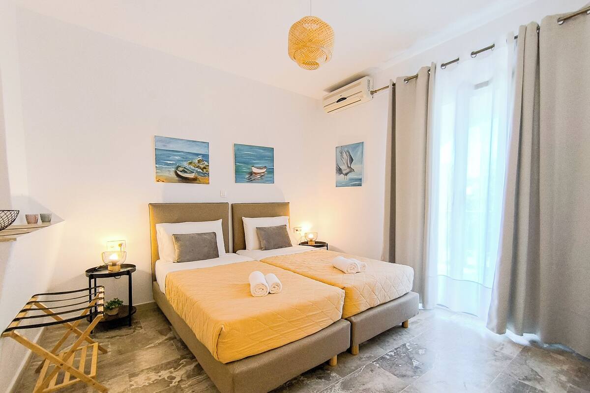 A double bed with a beige bedspread, two bedside tables with lamps, a wall-mounted air conditioner, and a window with curtains.