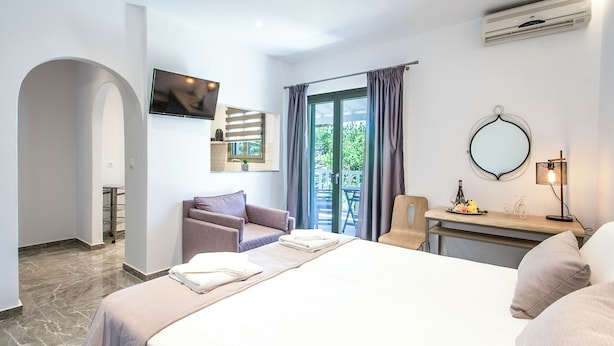 Ein modernes Hotelzimmer mit einem Bett, einer Sitzecke mit Sessel und Sofa, einem Schreibtisch mit Lampe sowie einem großen Fenster mit Vorhängen.