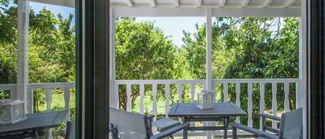 A covered outdoor patio with a table and chairs, surrounded by white railings and lush greenery.