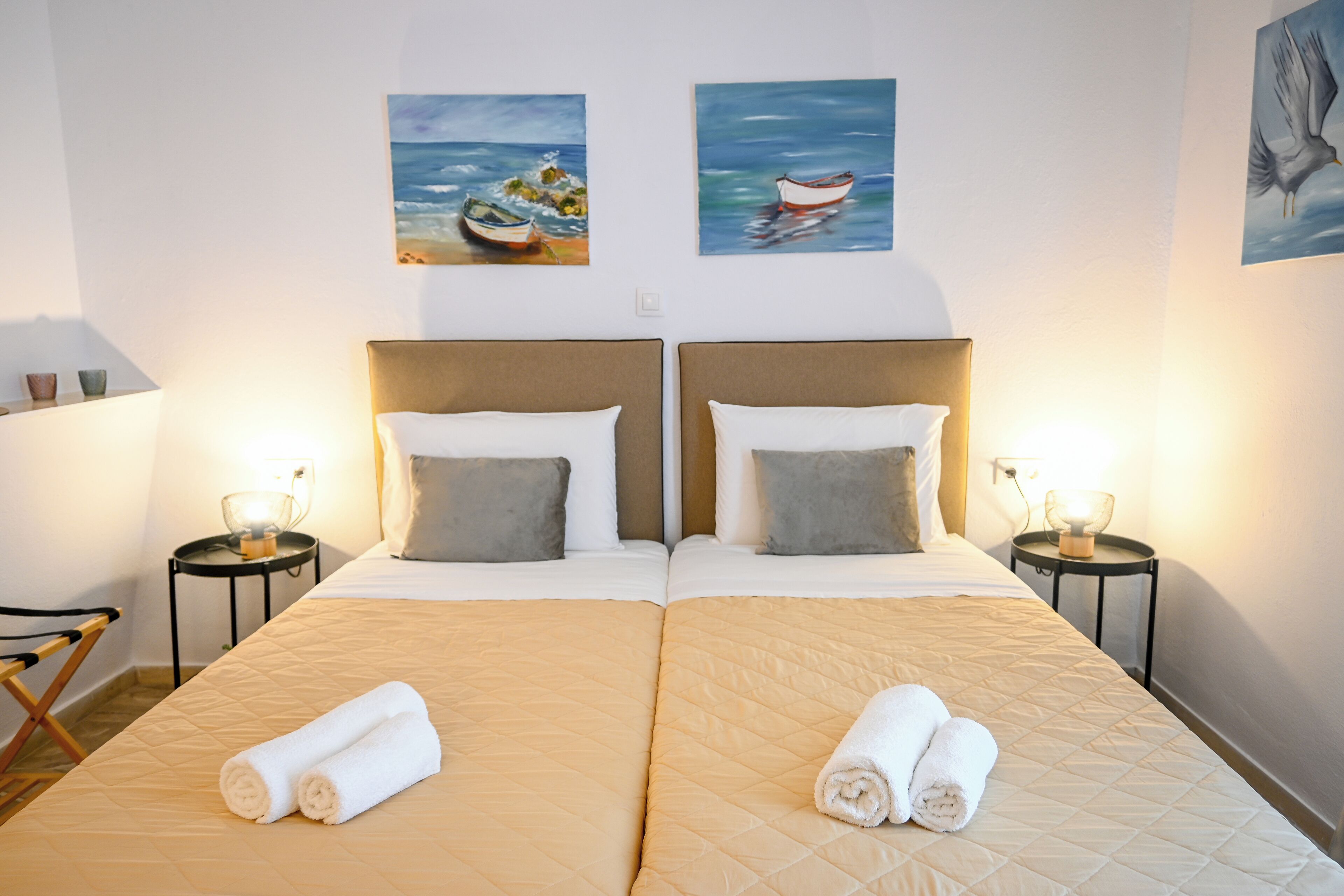 A double bed with a beige quilted cover, two pillows, and two rolled towels. There are two bedside tables with lamps, and artwork depicting boats on the wall.