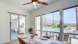Un coin repas avec une table dressée pour quatre personnes, de grandes fenêtres offrant une vue sur les montagnes et une piscine, ainsi qu’un ventilateur de plafond.
