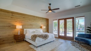 Une chambre avec un mur en bois, un lit avec une literie blanche, deux lampes de chevet, un ventilateur de plafond et de grandes fenêtres.