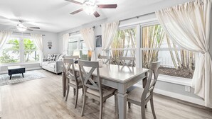 Un coin repas avec une table et des chaises en bois, un ventilateur de plafond et de grandes fenêtres avec des rideaux.