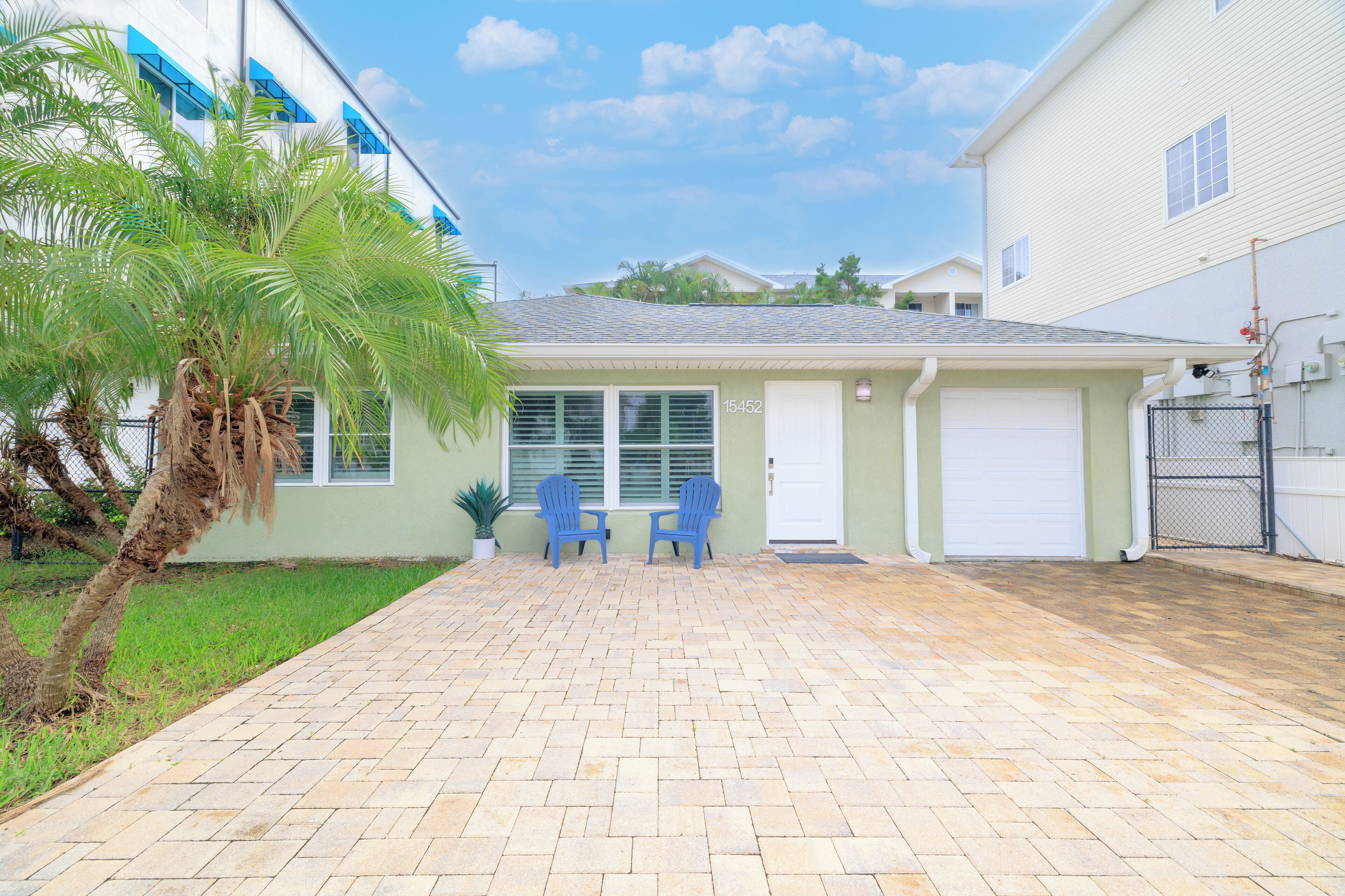 Una casa residencial con entrada pavimentada, dos sillas azules, una palmera y un garaje.
