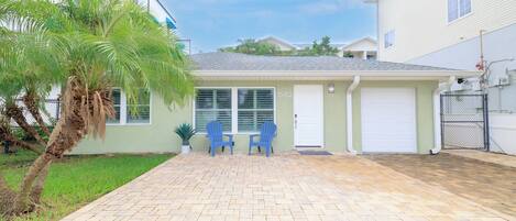 Una casa residencial con entrada pavimentada, dos sillas azules, una palmera y un garaje.