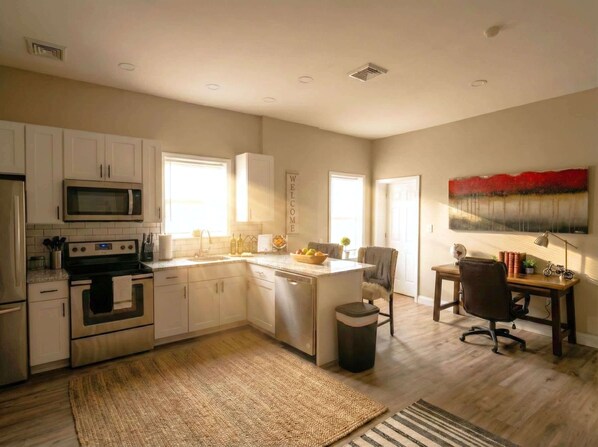 A modern kitchen with white cabinets, a microwave, an oven, and a sink. There's a dining area with a table and chairs, and a painting on the wall. - Charming Wilmington Penthouse Loft (Wilmington)