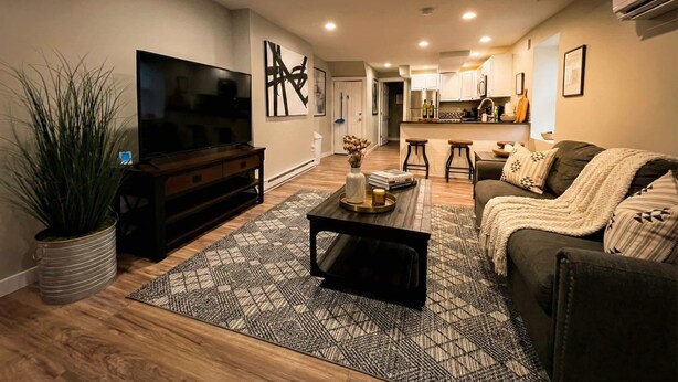 A modern living room with a flat-screen TV, a dark wooden entertainment unit, a patterned rug, a sofa with a throw blanket, and a kitchen area in the background. - Stylish Cool Springs Apt, King Bed (Wilmington)