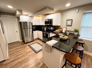 A modern kitchen with stainless steel appliances, white cabinetry, a black countertop, and a wooden floor. - Stylish Cool Springs Apt, King Bed (Wilmington)