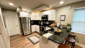 A modern kitchen with white cabinets, stainless steel appliances, a black countertop, and wooden stools. - Stylish Cool Springs Apt, King Bed (Wilmington)