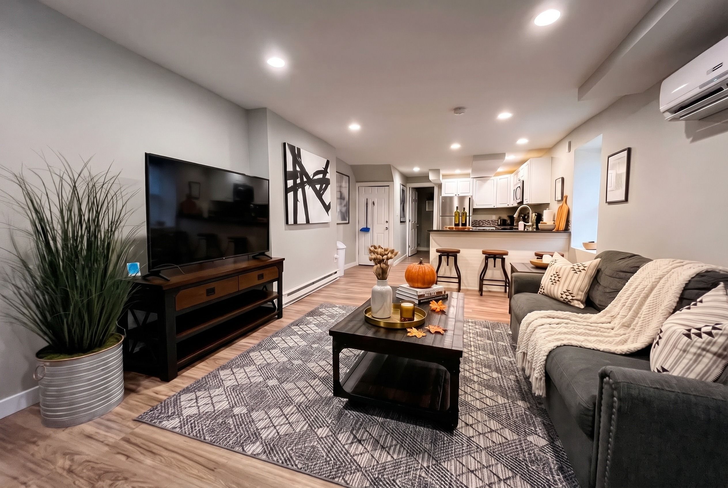 Ein modernes Wohnzimmer mit einem Flachbildfernseher, einer Unterhaltungsanlage aus dunklem Holz, einem gemusterten Teppich, einem grauen Sofa und einer Küche im Hintergrund.