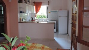 A kitchen with white cabinets, a refrigerator, a window with a curtain, and a dining area with a table and chairs. - Villa Sophia by CorfuEscapes (Corfu)