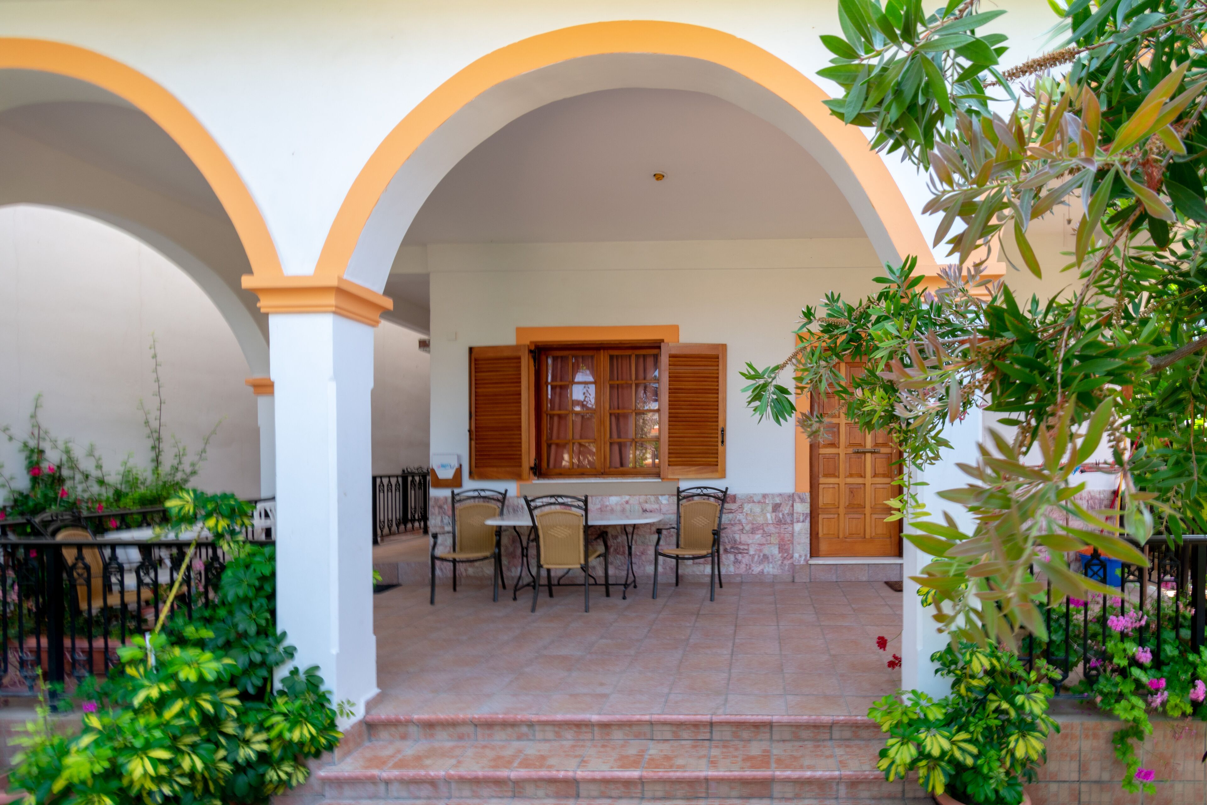A covered outdoor patio with a table and chairs, surrounded by arches and potted plants.