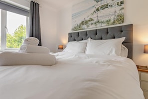 A neatly made bed with white linens and a headboard, a window with curtains, and a wall-mounted painting of a beach scene.