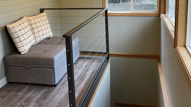 A room with a wooden ceiling, a ceiling fan, a window with a view of a house, a grey sofa with patterned cushions, and a staircase with a metal railing.