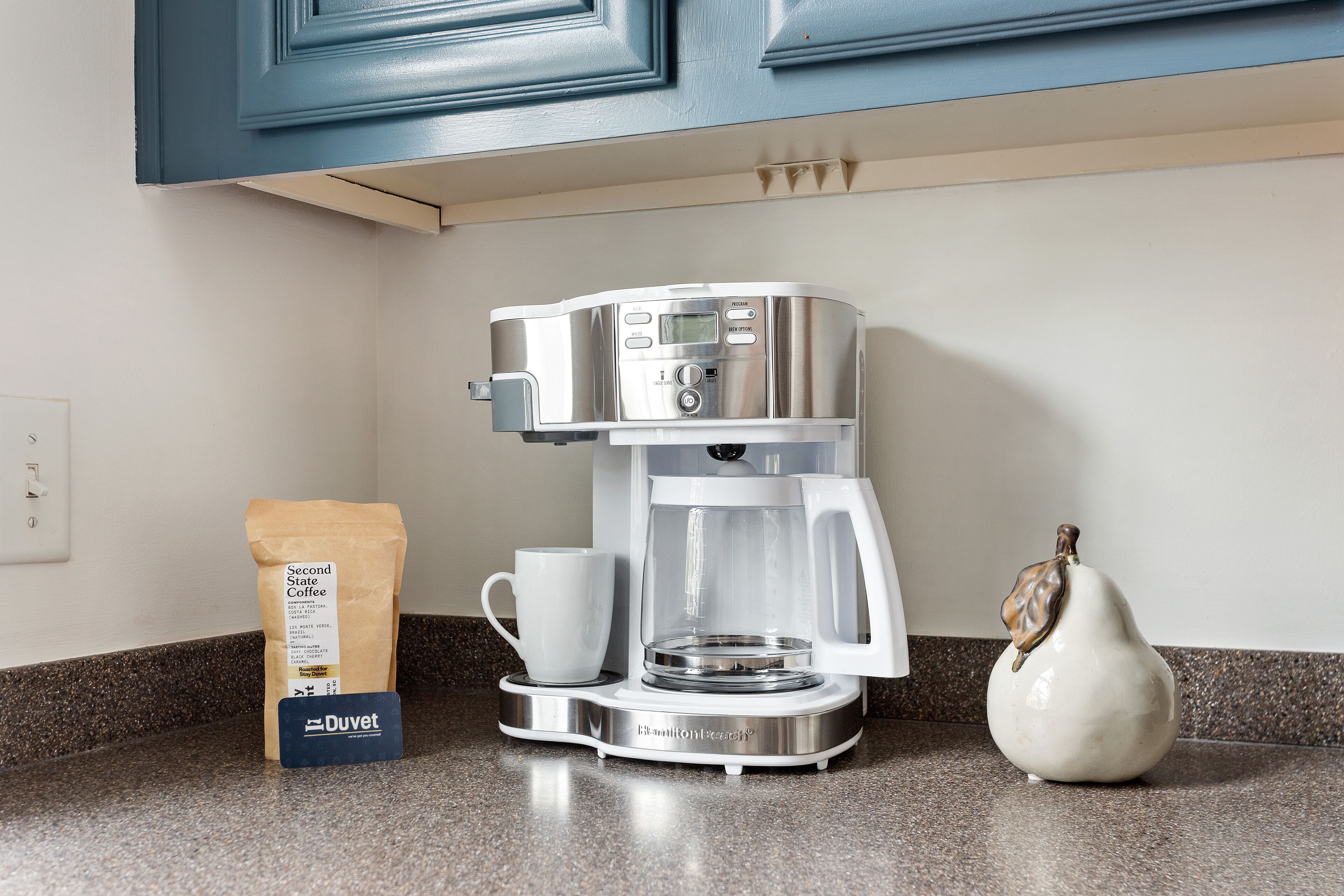A coffee maker on a countertop with a coffee bag, a mug, and a decorative item.