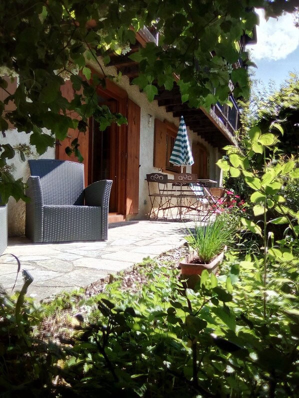 Eine Terrasse mit Korbmöbeln, einem Tisch und Stühlen, umgeben von Grünpflanzen und einem Blumentopf.
